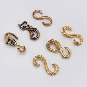 May include: A collection of brass-colored S-shaped hooks and decorative clasps. Designs include a skull, ornate scrollwork, and textured patterns. The hooks are displayed against a white background.