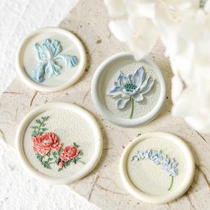 Op de afbeelding: Vier witte, cirkelvormige decoratieve stukken met verhoogde bloemmotieven. De bloemen zijn onder meer een iris, een lotus, twee pioenen en een tros kleine blauwe bloemen. De stukken zijn gerangschikt op een getextureerd, gebroken witte oppervlak.