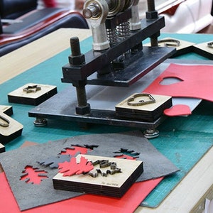 May include: A metal press with various wooden die-cut shapes and red and gray leather pieces. The shapes include stars, rectangles, and other designs. The scene is set on a wooden table with a green cutting mat.