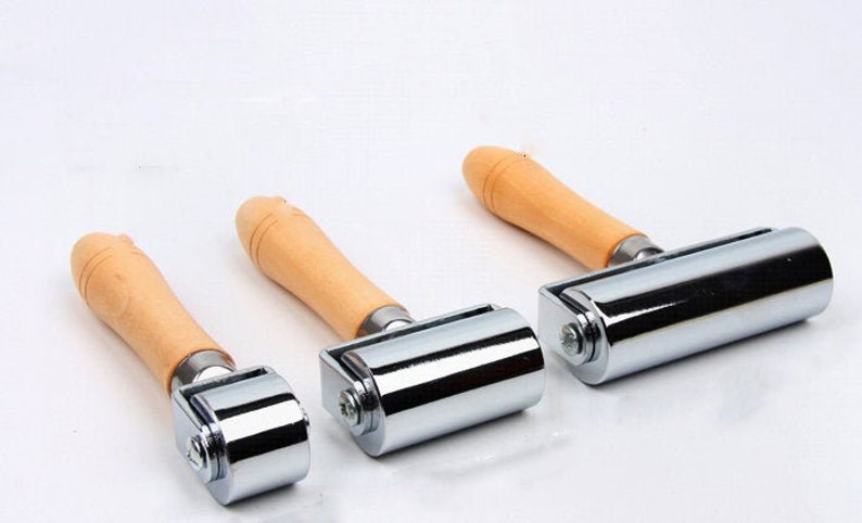 Peut inclure: Trois rouleaux &agrave; main en m&eacute;tal avec des manches en bois. Les rouleaux sont chrom&eacute;s et cylindriques, avec un petit boulon de chaque c&ocirc;t&eacute;. Les manches sont en bois clair, et les rouleaux sont dispos&eacute;s sur un fond blanc.