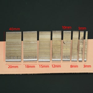 May include: A set of seven metal leather working tools, ranging in width from 3mm to 20mm, and 40mm in height. The tools are arranged on a tan leather strip against a dark background. The tools are used for cutting leather.