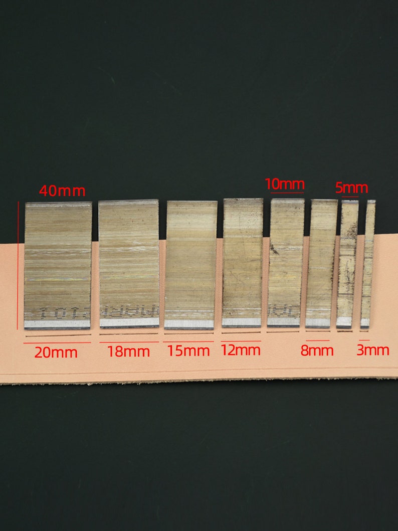May include: A set of seven metal leather working tools, ranging in width from 3mm to 20mm, and 40mm in height. The tools are arranged on a tan leather strip against a dark background. The tools are used for cutting leather.