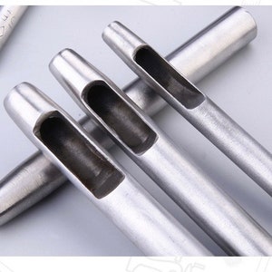 May include: Close-up of several silver-colored leather hole punches. The punches have a cylindrical shape with a rectangular opening at the end. One punch is partially visible in the background. The tools are designed for leathercraft and similar projects.