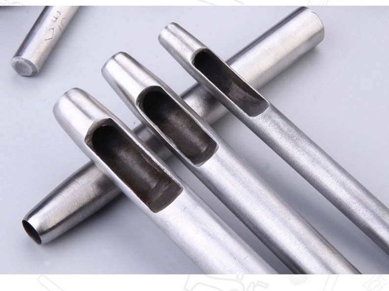 May include: Close-up of several silver-colored leather hole punches. The punches have a cylindrical shape with a rectangular opening at the end. One punch is partially visible in the background. The tools are designed for leathercraft and similar projects.