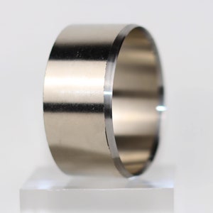May include: A silver-colored, cylindrical ring with a polished finish. The ring is made of a solid metal and has a smooth, reflective surface. The edges are cleanly cut, and the ring is displayed on a clear, rectangular stand.