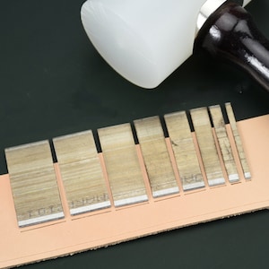 May include: A white mallet with a dark wooden handle rests above a series of leatherworking tools. The tools are arranged on a piece of tan leather, with a dark green background. The tools are rectangular with a silver edge and text.