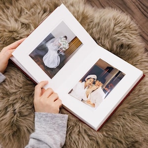 May include: A red-covered photo album with white mounts, open to reveal two photographs. One shows a bride with a bouquet, the other a couple in white robes. The album rests on a textured, brown surface.