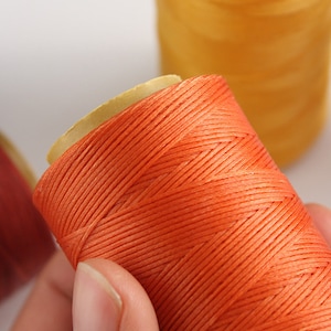 May include: Close-up of orange thread spools. The thread is tightly wound around a cardboard core. A second spool of yellow thread is visible in the background. The thread appears to be a vibrant, solid color.