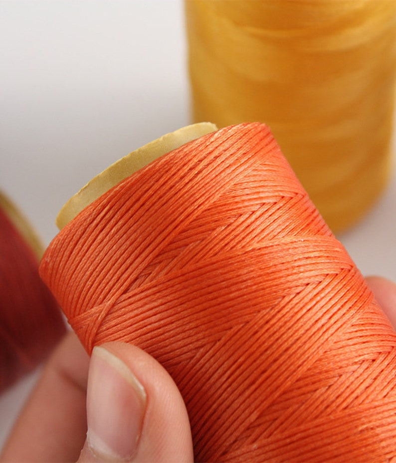 May include: Close-up of orange thread spools. The thread is tightly wound around a cardboard core. A second spool of yellow thread is visible in the background. The thread appears to be a vibrant, solid color.