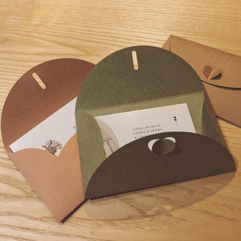 May include: Three heart-shaped paper envelopes in shades of brown and green, with a small heart cut-out and a slot at the top. The envelopes are open, revealing white cards with text and a floral illustration.