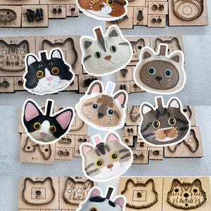 Puede incluir: Una colección de piezas de artesanía en cuero con forma de gato y troqueles de corte de madera. Las piezas de cuero presentan varios diseños de caras de gato en colores como marrón, negro y gris. Los troqueles de madera se utilizan para cortar el cuero en formas de gato.