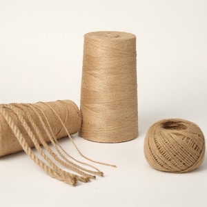 May include: A collection of natural brown jute twine in various forms. The image shows a large spool, a smaller ball, and a length of twine unraveled from a roll. The twine is tightly wound and has a rough texture, suitable for crafts and packaging.