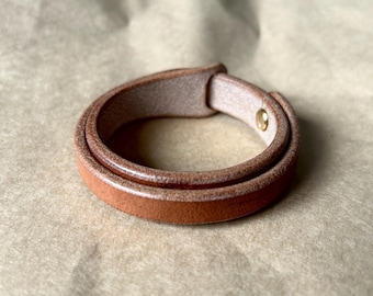 Może przedstawiać: Bransoletka ze skóry w kolorze brązowym, z podwójnym owinięciem. Bransoletka ma jasnobrązowe wnętrze i złote zapięcie na zatrzask. Skóra ma gładką fakturę i delikatny połysk. Szerokość: około 1,5 cm.