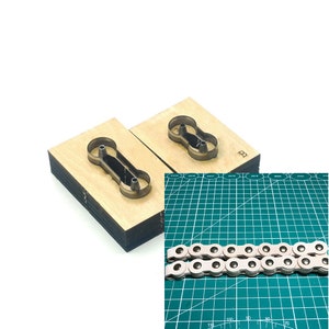 Op de afbeelding: Twee houten stansblokken met een kettingschakelontwerp, naast een gedeelte van een zilveren fietsketting. De blokken zijn lichtbruin met een donkere metalen omtrek van de kettingschakel. De ketting is zilverkleurig met witte leren riemen.