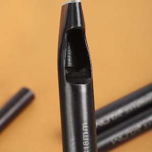 May include: Close-up of a black leather hole punch tool. The tool has a cylindrical body with a hollow, rectangular cutting end. The text "5x18mm" is printed on the side. The background is a warm, neutral color.