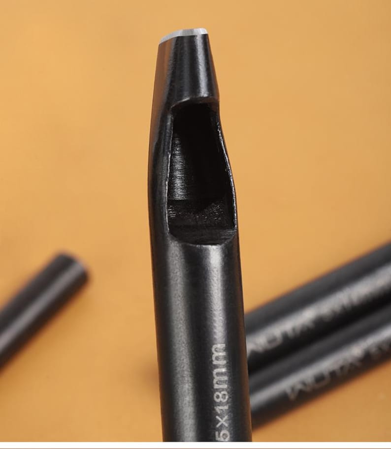 May include: Close-up of a black leather hole punch tool. The tool has a cylindrical body with a hollow, rectangular cutting end. The text "5x18mm" is printed on the side. The background is a warm, neutral color.