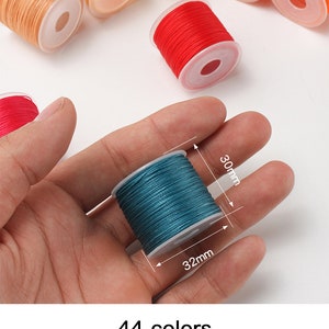 May include: A hand holding a teal spool of thread, 30mm high and 32mm wide. Other spools of thread in various colors, including orange, red, and tan, are scattered in the background. The image includes text indicating 44 colors, a line width of 0.5mm, and 20m/reel.