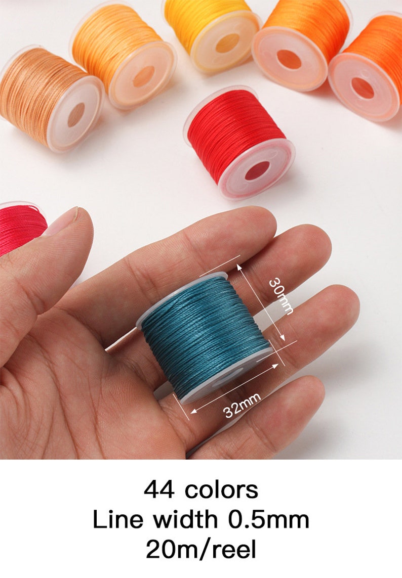 May include: A hand holding a teal spool of thread, 30mm high and 32mm wide. Other spools of thread in various colors, including orange, red, and tan, are scattered in the background. The image includes text indicating 44 colors, a line width of 0.5mm, and 20m/reel.