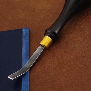 Può includere: Primo piano di uno strumento per la lavorazione della pelle con un manico in legno nero e una lama argentata curva. Lo strumento ha una fascia metallica color oro. Un taccuino blu con bordo blu è sullo sfondo. Lo sfondo è marrone.