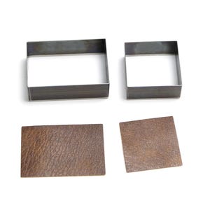 May include: Two metal square cutters and two pieces of brown leather. One cutter is rectangular, the other is square. The leather pieces are a rectangle and a square, both with a textured surface. The items are on a white background.
