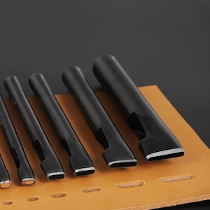 May include: A set of black leather working tools with various sized rectangular and oval shaped punches. The tools are arranged on a tan leather surface with pre-punched holes, against a dark background.