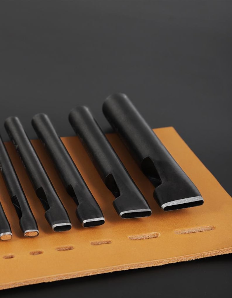 May include: A set of black leather working tools with various sized rectangular and oval shaped punches. The tools are arranged on a tan leather surface with pre-punched holes, against a dark background.