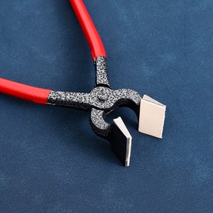 May include: Close-up of metal nipping pliers with red handles. The pliers are open, holding a small, light-coloured square object. The tool has a textured, dark grey finish.
