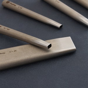 May include: Close-up of several metal chisels with square and rounded tips. The chisels are arranged on a dark surface, with the text "4/61" and "5 * 25" visible on the tools. The tools have a metallic finish.