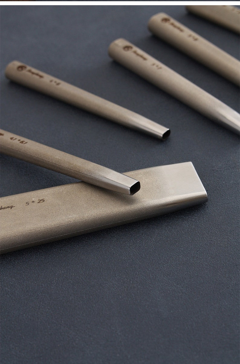 May include: Close-up of several metal chisels with square and rounded tips. The chisels are arranged on a dark surface, with the text "4/61" and "5 * 25" visible on the tools. The tools have a metallic finish.