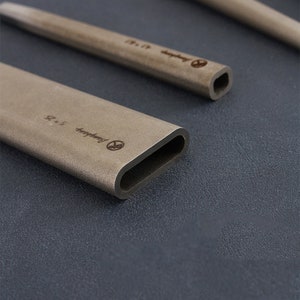 May include: Several light brown, rectangular, hollow tubes with rounded edges are displayed on a dark grey surface. Each tube has engraved text and a logo. The dimensions are 5 x 2.5. The tubes are made of a matte material.