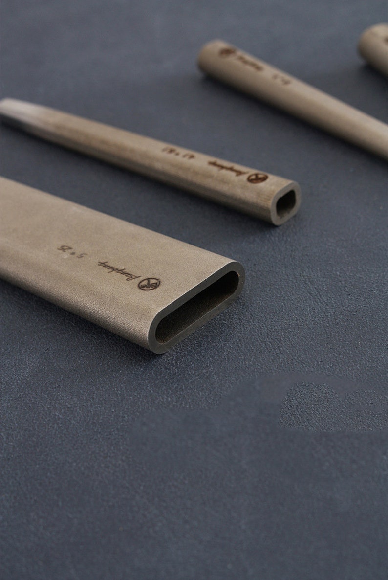 May include: Several light brown, rectangular, hollow tubes with rounded edges are displayed on a dark grey surface. Each tube has engraved text and a logo. The dimensions are 5 x 2.5. The tubes are made of a matte material.