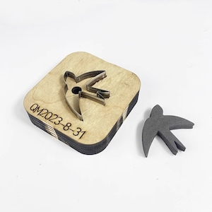 May include: A wooden die-cut tool with a metal outline of a bird shape, sitting on a stack of wooden squares. A separate gray bird-shaped foam cutout is next to the die-cut. The text "QM2023-8-31" is visible on the wooden square.