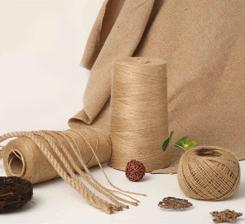 May include: An assortment of natural jute twine and rope in various forms. The image shows a large spool, a smaller roll, and a ball of twine, along with decorative woven spheres and burlap fabric in the background.