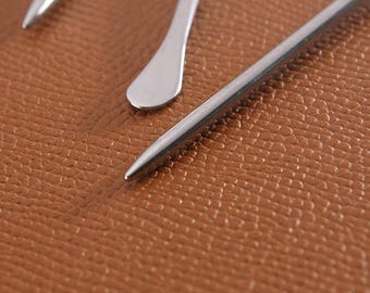 May include: Close-up of three stainless steel tools with pointed and rounded ends. The tools are arranged diagonally against a textured, brown leather surface. The tools have a shiny, metallic finish.