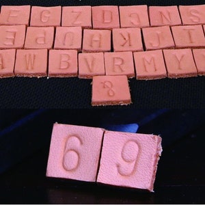 Puede incluir: Una colecci&oacute;n de peque&ntilde;as baldosas cuadradas de cuero bronceado con letras y n&uacute;meros en relieve. La secci&oacute;n superior muestra el alfabeto, mientras que la inferior muestra los n&uacute;meros 6 y 9. Las baldosas est&aacute;n dispuestas sobre una superficie oscura.