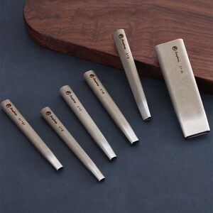 May include: A collection of silver metal punches in different sizes, displayed on a dark blue surface. Each punch has a rectangular end and is marked with its size. A rectangular metal holder is also present.