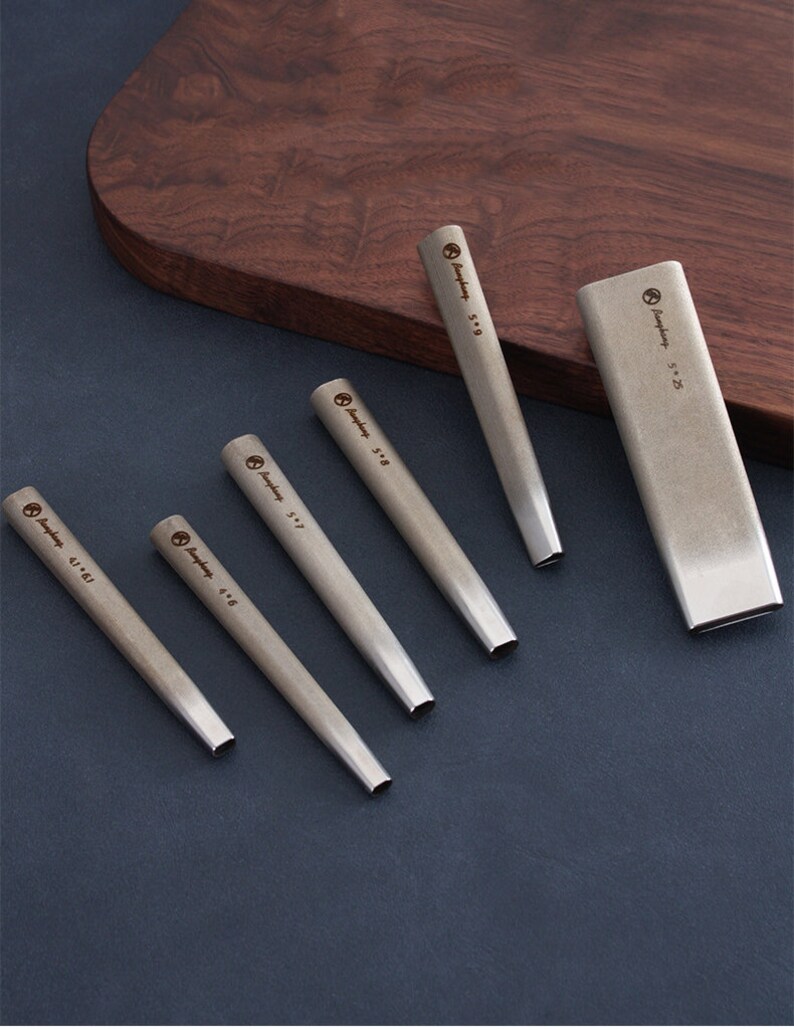 May include: A collection of silver metal punches in different sizes, displayed on a dark blue surface. Each punch has a rectangular end and is marked with its size. A rectangular metal holder is also present.