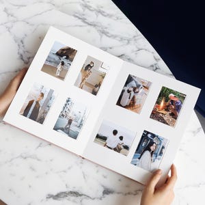 Peut inclure: Album photo ouvert présentant plusieurs photographies. L'album contient diverses petites photos de personnes, de chats et de scènes en extérieur. L'album est tenu ouvert par des mains et repose sur une surface en marbre.