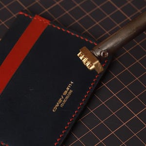 May include: A navy blue leather cardholder with a red stripe and red stitching. The cardholder features the text "Crazy Smith Handmade" in gold lettering. A metal branding tool is positioned above the cardholder.