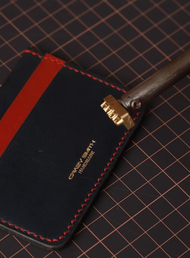 May include: A navy blue leather cardholder with a red stripe and red stitching. The cardholder features the text "Crazy Smith Handmade" in gold lettering. A metal branding tool is positioned above the cardholder.