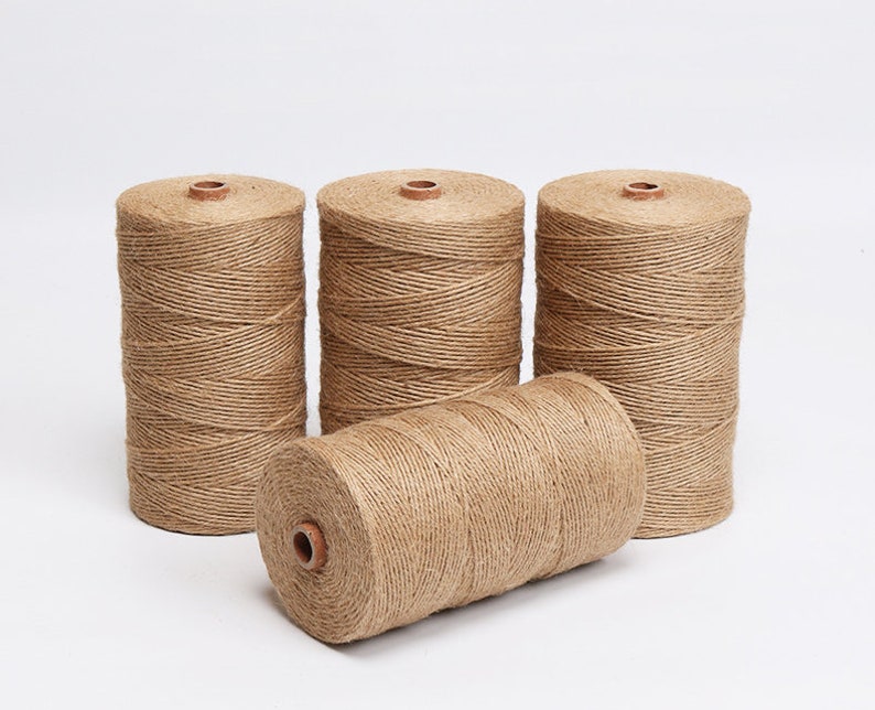 May include: Four spools of natural-colored twine. Three spools stand upright, while one lies on its side. The twine is tightly wound around a central core, creating a cylindrical shape. The spools are set against a plain white background.