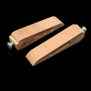 May include: Two wooden leathercraft clamps with a natural finish. Each clamp has a curved top, a recessed groove, and a metal screw for adjusting the grip. The clamps are designed for holding leather pieces together during crafting.