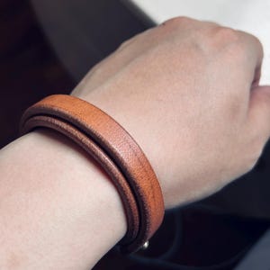 May include: A double-wrap leather bracelet in a warm, medium brown hue. The bracelet is wrapped around a wrist, showcasing its smooth texture and simple design. The bracelet appears to be approximately 0.5 inches wide.