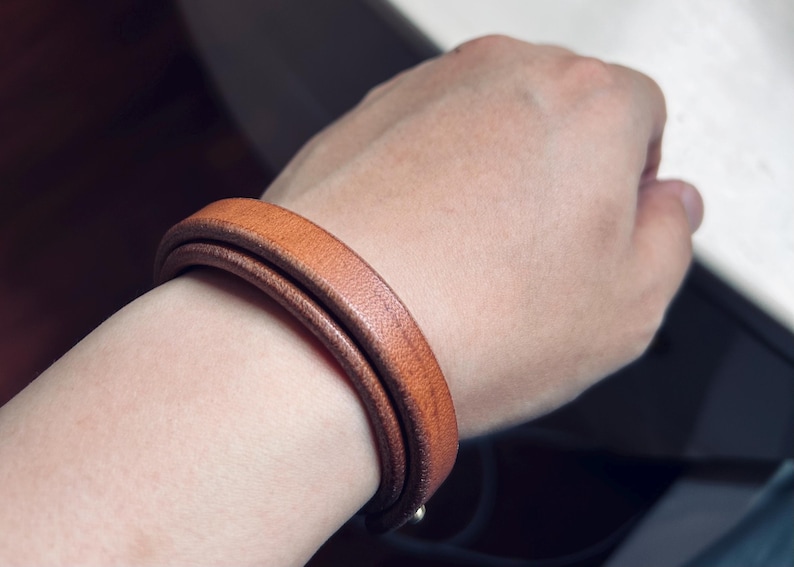 May include: A double-wrap leather bracelet in a warm, medium brown hue. The bracelet is wrapped around a wrist, showcasing its smooth texture and simple design. The bracelet appears to be approximately 0.5 inches wide.
