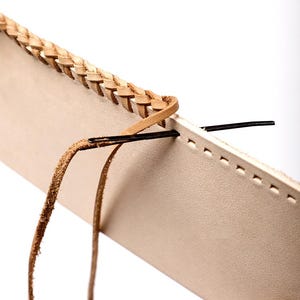 May include: Close-up of a leather craft project in progress. The image shows a light tan leather piece with a braided edge. A needle and thread are inserted through the leather, indicating hand stitching. The background is plain white.