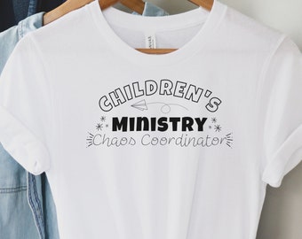 May include: White t-shirt with the text "CHILDREN'S MINISTRY Chaos Coordinator" in black. The design includes a stylized airplane and starbursts. The shirt is hanging on a wooden hanger with other clothing items in the background.