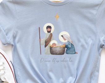 May include: Light blue t-shirt featuring a nativity scene illustration with Joseph, Mary, and baby Jesus. The design includes a star and the text "O come, let us adore him." White sneakers and sunglasses are visible.