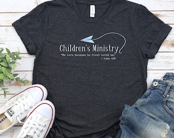 May include: Dark gray t-shirt with the words "Children's Ministry" and the quote "We love because he first loved us." 1 John 4:19. The design includes a blue and white paper airplane graphic. White sneakers and blue jeans are also in the image.