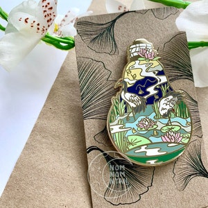 Puede incluir: Un pin de esmalte dorado con una ilustración estilizada de una botella de vidrio con un paisaje azul, verde y blanco en su interior. El paisaje incluye dos grullas blancas, nenúfares y una luna. El pin está sobre un fondo marrón con un diseño de hoja de ginkgo.