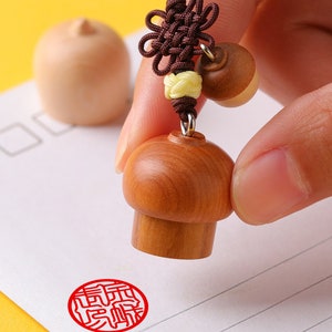 May include: A wooden stamp with a mushroom-shaped handle and a red ink pad. The stamp is being used to create a red impression of a Chinese character on a white piece of paper.